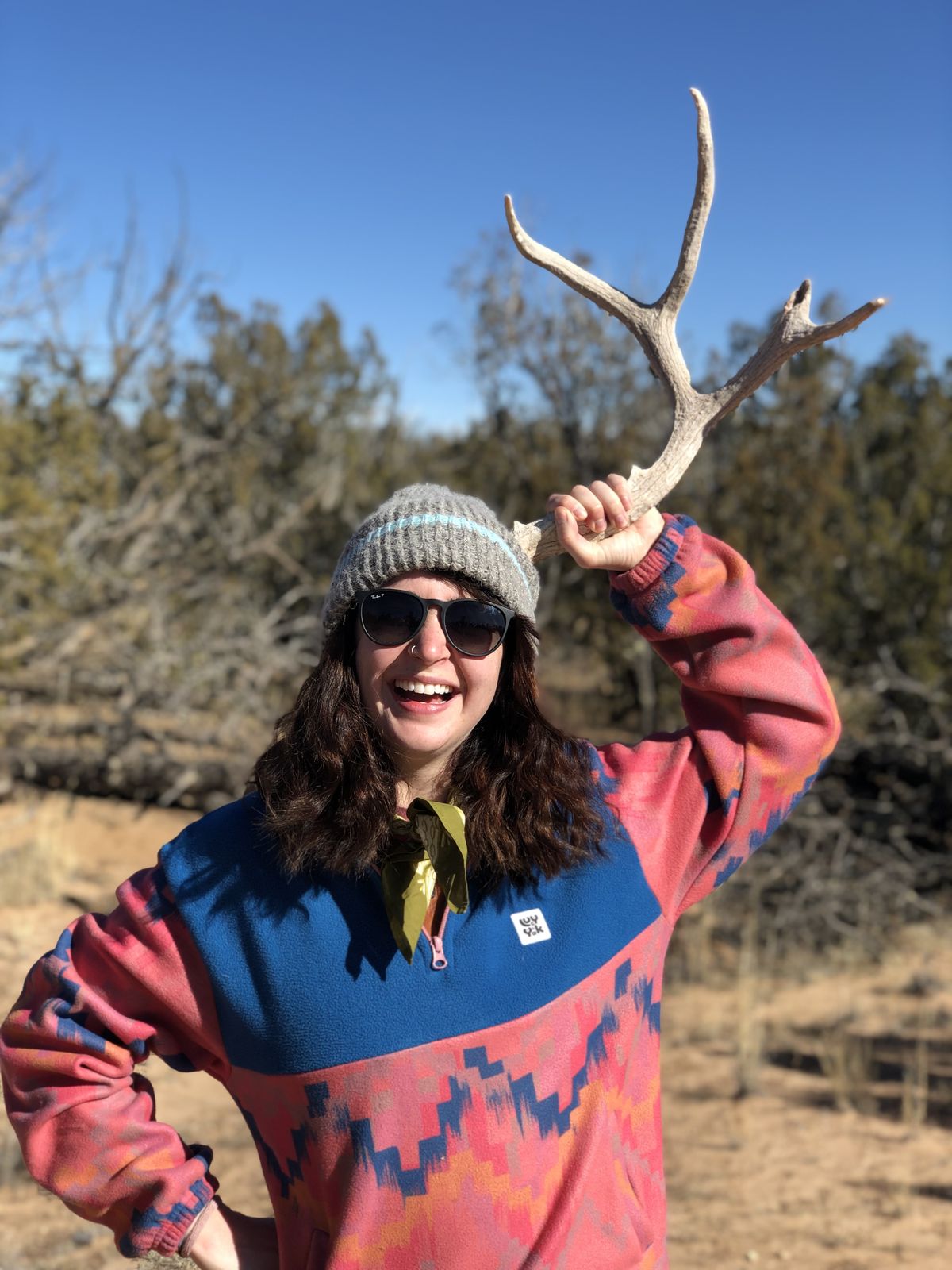 Leigh-Anne laughing in sunglasses and a beanie, holding a deer antler over her head in the desert